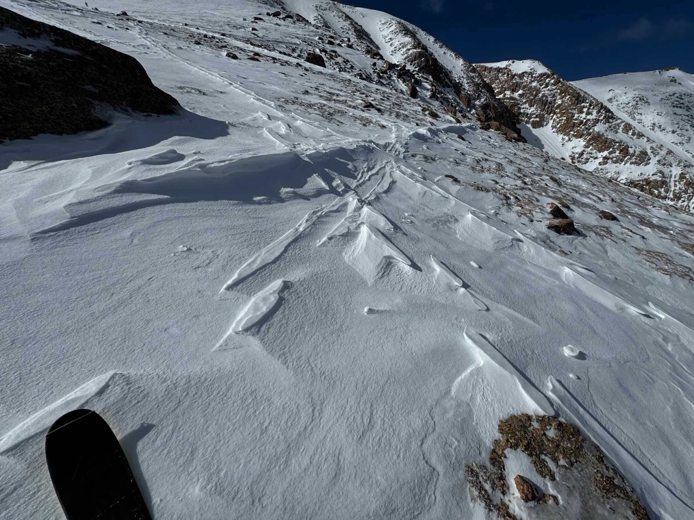 Wind-scoured snow on a high alpine ridge, with a ski tip in the foreground.
